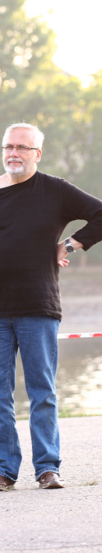 A man with gray hair and a beard stands outdoors in front of a river during Kapusvētki, wearing a black long-sleeve shirt and blue jeans. He holds a professional video camera in his right hand and has his left hand on his hip. The background includes trees, a building, and a red-and-white striped barrier.