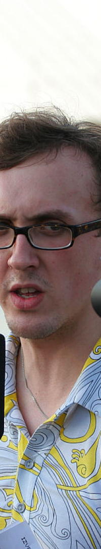 A man in glasses, wearing a white and yellow patterned shirt, speaks into a microphone in front of a video camera during the Oligarhu kapusvētki. Another man stands to his left, wearing a black shirt with a logo. In the background, a river and a bridge are visible under a cloudy sky.
