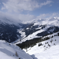 A panoramic view of a snowy mountain landscape in the Austrian Alps. Snow-covered peaks and valleys stretch across the scene under a partly cloudy sky. Evergreen trees dot the slopes, and a narrow valley cuts through the center. The snow sparkles in the sunlight, creating a serene winter wonderland.