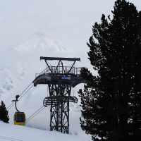 A yellow cable car ascends on a cable towards a metal support tower in the Austrian Alps. The scene is set in a snowy mountainous area with snow-covered peaks visible in the background. To the right of the tower, tall, dark evergreens provide contrast to the white landscape.