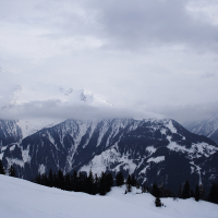 A vast snow-covered mountainous landscape under a cloudy sky. The Austrian Alps are partially obscured by low-hanging clouds, with patches of dark evergreen trees dotting the slopes. The foreground features a gentle snow-covered incline, while the background showcases rugged mountain peaks.