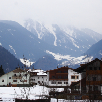 A serene, snow-covered village nestled in a mountainous region of the Austrian Alps is depicted. Traditional wooden houses with white facades are scattered amidst bare trees. The towering mountains in the background are partly shrouded in mist, and patches of snow can be seen on their slopes. The scene is tranquil and picturesque.