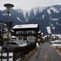 A narrow asphalt road lined with wooden fences runs through a snow-covered village in the Austrian Alps. Traditional alpine buildings with dark wooden roofs sit beside the road. A snow-capped mountain range is visible in the background under an overcast sky. A streetlamp and parked cars are on the left.