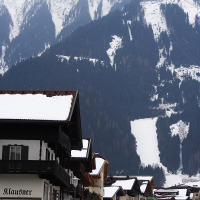 A snowy mountain landscape in the Austrian Alps boasts tall, forested peaks partially covered in clouds. Below, a quaint village features traditional alpine houses with sloped roofs. A sign reads "Klaushof" and "Parkplatz Sporthotel Mannl," indicating a parking area for a nearby sport hotel.