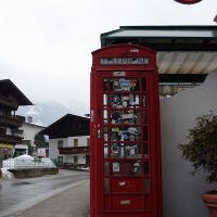 A traditional red British telephone booth stands on a wet street in a small town with alpine-style buildings, reminiscent of an Austrian village. The booth is adorned with stickers. In the upper right corner, a round Heineken beer sign is attached to a building. Snowy Alps are visible in the background.