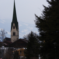 A tall church steeple with a green roof stands prominently in a snow-covered town, reminiscent of the Austrian Alps. Surrounding buildings are nestled among leafless trees, and several benches are visible in the foreground. The sky is overcast, adding a somber tone to the winter scene.
