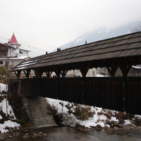 A covered wooden pedestrian bridge spans over a narrow stream in a snowy landscape. A stone path leads to the bridge, and snow patches are visible on the ground and rooftops. In the background, a church with a red steeple stands among buildings and trees, set against the misty Austrian Alps.