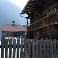 A rustic wooden house with a slanted roof and a balcony is set against the majestic Austrian Alps. A weathered wooden fence is in the foreground, and a second wooden building with a chimney is visible behind. The scene appears serene and misty, suggesting a cold, possibly winter day in Austria.