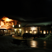A cozy restaurant named "Mittergrat" in the snow-covered Austrian Alps is illuminated at night. The building features large windows and warm lights, casting a soft glow on the snowy ground and parked cars. A streetlight provides additional lighting, and snow piles can be seen in the foreground.