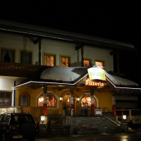 A two-story building with a lighted "Pizzeria" sign hangs above an entrance adorned with string lights. Snow blankets the roof, reminiscent of the Austrian Alps. Three parked cars are in front. The facade has various light sources, creating a warm, inviting glow against the night sky. Another sign on the left of the building reads "Nah & Frisch".