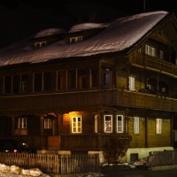 A wooden alpine house with a snow-covered roof at night, nestled in the Austrian Alps. Warm yellow lights glow through some windows. The house has a balcony with ornate wooden railings. A fence surrounds the property; a car is partially visible on the left side near the entrance amidst the snow-covered ground.