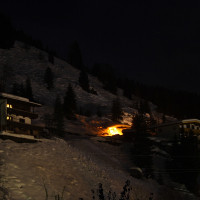 Nighttime scene of a snowy mountainside in the Austrian Alps with two houses. One house is brightly illuminated, with a fire burning in front. A small, dimly lit structure is nearby. Trees and patches of snow are visible on the slope, and the sky is dark, creating a contrast with the lights and fire.
