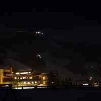 A serene nighttime scene of a snow-covered village nestled in the Austrian Alps. Several buildings are warmly lit from within, casting a cozy glow against the dark, snow-laden slopes. Lights speckle the distant hills, and stars can faintly be seen in the clear night sky.