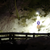 A snowy scene at night in the Austrian Alps features a wooden railing partially visible in the foreground. A lamppost illuminates a pedestrian path sign and a "no vehicles over 3.5 tons" sign. Leafless trees and snow-covered ground surround the area, with the lamppost casting a warm light on the snow.