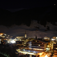 A nighttime view of a picturesque, snow-covered village nestled in the Austrian Alps valley, illuminated by warm yellow lights. The village features numerous small buildings, a prominent church with a tall steeple, and hills covered in snow and pine trees in the background. A crescent moon is visible in the dark sky.