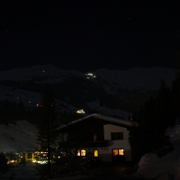 A serene nighttime mountain village scene in the Austrian Alps. Dim lights from houses illuminate the snowy landscape. A dark wooden chalet is in the foreground with warm lights in the windows. Snow-covered slopes and tall trees are set against a starry sky, with distant peaks barely visible in the dark.