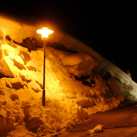 A snow-covered path in the Austrian Alps is illuminated by old-fashioned streetlights glowing with warm yellow light against a dark night sky. The snow forms high banks on either side of the narrow pathway, creating a serene winter scene. There is a person walking in the distance, partially visible beyond the curve.