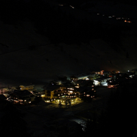 A nighttime view of a small, illuminated village nestled in the Austrian Alps. The buildings are lit with warm, yellow lights, creating a cozy atmosphere amidst the surrounding darkness. Snow-covered hills and trees are faintly visible in the background, hinting at a winter setting.