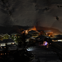 A nighttime aerial view of a snowy alpine village in the Austrian Alps, illuminated by warm yellow lights from the windows and street lamps. The snow-covered rooftops and surrounding snow-covered landscape create a serene, wintry atmosphere. Tracks in the snow are visible in the background on the hillside.