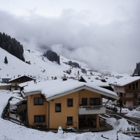 A snow-covered mountain village with several beige and brown chalets lies nestled in the Austrian Alps. The rooftops are blanketed in snow, and a winding path passes through the village. Tall evergreen trees surround the area, and fog partially obscures the mountainous background. The scene exudes winter tranquility.