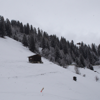 A snowy mountain landscape in the Austrian Alps, with a few small wooden huts scattered on a hillside. The hillside is covered in snow with sparse evergreen trees on the upper half. A windsock is visible near the bottom of the image. The overcast sky adds to the serene and quiet atmosphere.