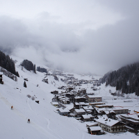 A snow-covered village nestled in a valley surrounded by the dense, evergreen forests and mountains of the Austrian Alps. The village features numerous buildings with snow-capped roofs. Skiers are visible on the slopes, while mist and clouds partly obscure the mountains in the background, adding a serene, wintry atmosphere.