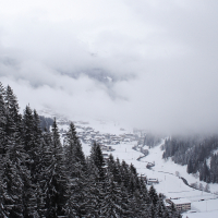 A snow-covered village nestled in a valley surrounded by dense pine forests and blanketed in mist and clouds lies within the Austrian Alps. Buildings are sprinkled throughout the landscape, and a winding road weaves through the snowy terrain. Tall trees in the foreground frame the serene, wintery scene.