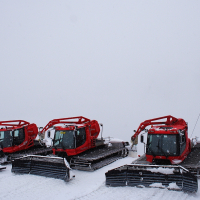 Three red snow groomers are lined up on a snowy slope in the Austrian Alps, with a grey ski lift tower and gondolas on the right. The vehicles have large, black, toothed plows in front and are covered in a light layer of snow. The sky is overcast, blending seamlessly with the white, snow-covered ground.