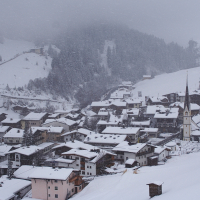 A quaint village nestled in the snowy Austrian Alps. Snow-covered houses are densely packed on a sloping hill, with a prominent church and tall steeple in the foreground. The background features snow-dusted evergreen trees and a foggy, overcast sky, evoking a serene winter atmosphere.