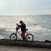 A young girl stands beside her bicycle on a rocky shore near the Daugavgrīva lighthouse, facing the ocean. She is wearing a purple hoodie and red pants. The ocean waves gently crash against the rocks, and the sky above is overcast with soft light filtering through the clouds. The beach stretches in the foreground.