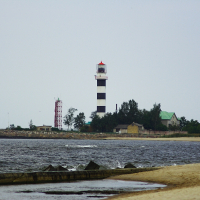 A coastal scene with the Daugavgrīva black and white striped lighthouse standing tall near the shoreline. Adjacent to the lighthouse are small buildings with green roofs. There is a red communication tower nearby. The beach has a gentle curve, with waves approaching the sandy shore under a cloudy sky.