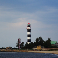 A black and white striped lighthouse stands on the coastal shore of Daugavgrīva under a cloudy sky. Surrounding the lighthouse are a few small buildings, including one with a green roof. The foreground has a sandy beach and calm ocean waves lapping the shore. Trees and a red and white tower are adjacent to the lighthouse.