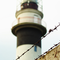 A tall lighthouse with black and white horizontal stripes stands slightly out of focus in the background. Known as the Daugavgrīva lighthouse, it features a lantern room at the top with protective railings. In the foreground, a rusty barb wire fence is sharply in focus, with a weathered concrete wall beneath it.