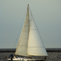 A lone sailboat with white sails fully extended glides on a calm body of water near the Daugavgrīva lighthouse. The sky above is overcast, and a distant, dark horizon line separates the sky from the water. Two people are on the boat's deck, possibly navigating or adjusting the sails.