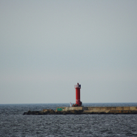 A red lighthouse stands at the end of a concrete pier jutting into a calm, expansive body of water under a clear, pale sky. A few people are gathered around the base of the Daugavgrīva lighthouse, and the horizon stretches out in the distance.