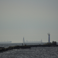 A hazy, overcast scene of a rocky shoreline extending into the sea with a small white lighthouse at Daugavgrīva and a few people near it. Several large ships and a sailboat are visible on the horizon under the gray sky. The water appears calm with gentle waves.