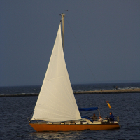 A single-mast sailboat with white sails and a yellow hull is sailing on calm blue waters near Daugavgrīva. Two people can be seen onboard under a blue canopy. The background features a narrow strip of land with a few people walking by the lighthouse, and the sky is a gradient from light to dark blue.