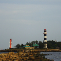 A rocky pier extends out into the water, leading to Daugavgrīva with two lighthouses. The near lighthouse is black and white, while the farther lighthouse is red and white. A small, green-roofed building stands between them, surrounded by trees. The sky is overcast.