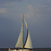 A white sailboat with intricate black designs on its hull glides through the water near the Daugavgrīva lighthouse. Two people are on board, one wearing a yellow jacket. The boat’s sail is fully set, catching the wind. The sky is overcast with shades of blue and gray, and the water appears calm.