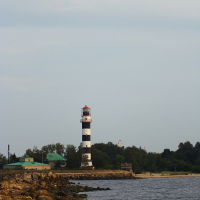 A striped lighthouse with black and white horizontal bands stands near a rocky shoreline, reminiscent of the Daugavgrīva lighthouse. Surrounding the scene are several small buildings and trees. The calm water of the sea extends towards the right, under a cloudy, light blue sky.