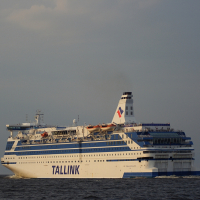 A large white passenger ferry with the name "TALLINK" in blue letters on the side sails past a distant lighthouse on the calm sea. The ship has several decks with visible lifeboats and is topped by a white smokestack with a blue and red logo. The sky above Daugavgrīva is mostly clear with a few clouds.