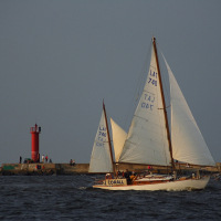A sailboat named "Corall" with sail number LAT 740 glides on dark waters near a rocky breakwater. A vivid red lighthouse at Daugavgrīva stands on the breakwater, contrasting with the muted sky and sea. Several people are visible near the lighthouse, and the scene is calm, suggesting a peaceful day.