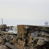 A stone pier extends into the sea, with large rocks scattered along its path. Near the end of the pier, Daugavgrīva lighthouse stands proudly. In the distance, a blue and white ferryboat sails on the horizon under an overcast sky. Two people are visible walking on the pier.