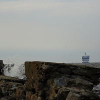 A rocky shoreline with large waves crashing against the rocks. A white lighthouse stands on the left side of the image near the edge of the shore, reminiscent of Daugavgrīva's historic beacon. In the distance, a large ferry can be seen on the horizon of the open sea under a cloudy, pale sky.
