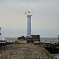 A solitary white Daugavgrīva lighthouse stands at the end of a weathered, concrete jetty extending into calm sea waters. To the left, a small sailboat with a white sail glides by, while a large ferry is visible in the distance on the right. The sky above is overcast with a pale, muted light.