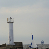 A white lighthouse stands at the edge of a concrete pier extending into the sea at Daugavgrīva. A sailboat with a white hull and blue sail is nearby in the water. In the distance, a large ferry or cruise ship is visible on the horizon under a cloudy sky. The scene exudes a calm, maritime atmosphere.