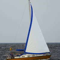 A yellow and white sailboat is sailing on a body of dark, choppy water, near the historic Daugavgrīva lighthouse. The sail is primarily white with a blue trim along the edges. There are a few people on board, wearing jackets. The sky is overcast, suggesting a cloudy day. A small yellow buoy is visible in the distance behind the sailboat.