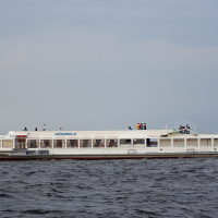 A white passenger ferry named "Jūrmala" is sailing on the waters of Daugavgrīva under a cloudy sky. The ferry, with its large windows, is flying a red and white flag at the stern. People on deck are enjoying the view, possibly spotting the distant lighthouse as they navigate slightly choppy waters.