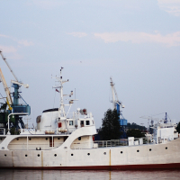 A vintage white ship docked at a harbor, featuring a rounded stern and multiple decks with various structures. Several cranes, painted yellow and blue, stand in the background near the Daugavgrīva lighthouse. The ship reflects a serene evening sky with a hint of pink, along with the calm water surface it is moored on.