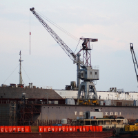 A large ship is docked for repairs in a Daugavgrīva shipyard. Two cranes are visible: one in the foreground lifting equipment onto the ship and another in the background. The lower part of the hull displays weathering marks. Red barriers with unreadable white text can be seen, and a distant lighthouse stands sentinel.
